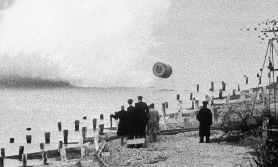 During a trial run demonstration, British officials watch the spinning mine bounce across the water toward the shoreline. Barnes Wallis is at far left, urging the bomb on. (IWM FLM 2343)