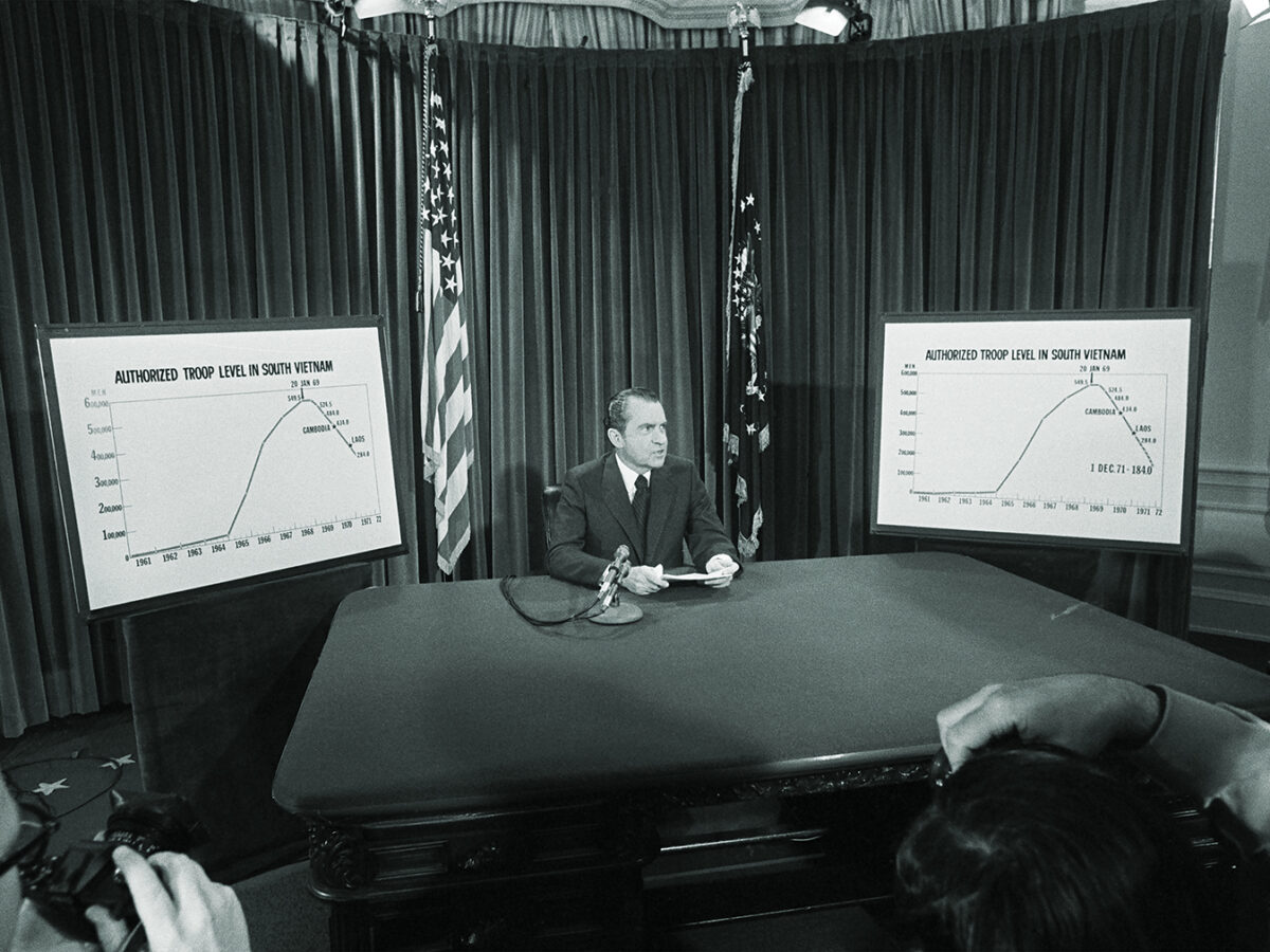 Photo of President Nixon flanked by charts he used to illustrate his televised speech from the White House 4/7 in which he announced he will withdraw an additional 100,000 U.S. troops by December 1. The charts show the authorized troops level in South Vietnam.