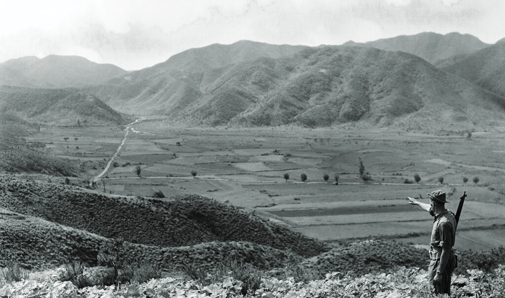 Photo of the ridge known as the “Hook” a rifleman of the 1st Battalion of the British Gloucestershire Regiment (aka “Glorious Glosters”) points across the valley where countless Chinese died.