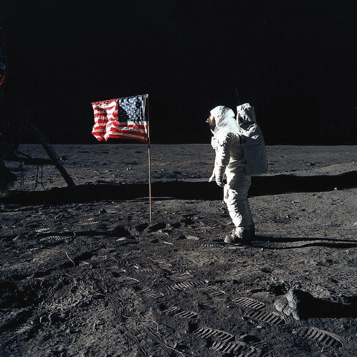 Photo of astronaut Edwin E. Aldrin Jr., lunar module pilot of the first lunar landing mission, poses for a photograph beside the deployed United States flag during an Apollo 11 extravehicular activity (EVA) on the lunar surface. The Lunar Module (LM) is on the left, and the footprints of the astronauts are clearly visible.